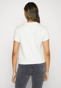 Hvit t-skjorte med korte ermer og rund hals, med en liten rød logo på siden, kombinert med mørkegrå jeans.