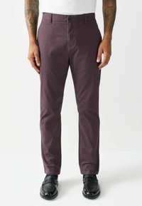 Burgunderfarbene, maßgeschneiderte Hose mit flacher Vorderseite, Gürtelschlaufen und Knopfverschluss; der Stoff hat eine glatte Textur. Getragen mit schwarzen Slippern.