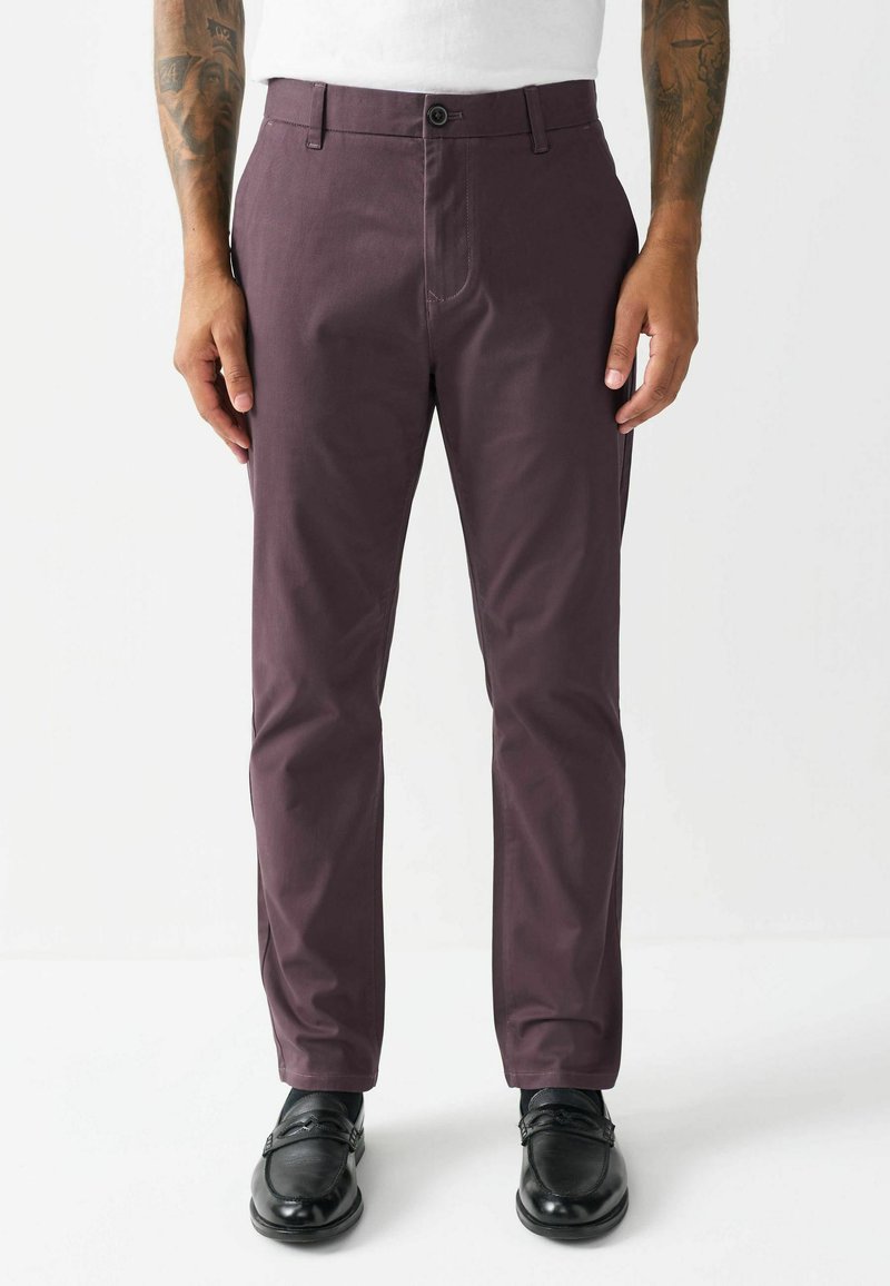 Burgunderfarbene, maßgeschneiderte Hose mit flacher Vorderseite, Gürtelschlaufen und Knopfverschluss; der Stoff hat eine glatte Textur. Getragen mit schwarzen Slippern.