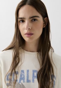 Lys beige sweatshirt med blå trykt tekst og design. Modellen har langt mørkebrunt hår og naturlig makeup, og står med front mod en ensfarvet baggrund.