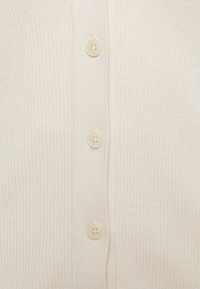 Krämfärgad ribbad kofta med knäppning fram, med fem ljusa plastknappar. Mjuk textur med figurnära passform.