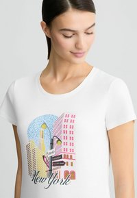 T-shirt en coton blanc avec un graphisme coloré représentant la ville de New York, avec des bâtiments en rose et jaune, ainsi que des panneaux de rue décoratifs.