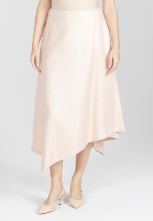 Kobieta ubrana w jasnoróżową asymetryczną spódnicę midi i beżowe sandały na obcasie z paskiem z tyłu, stojąca na jasnym, jednolitym tle.