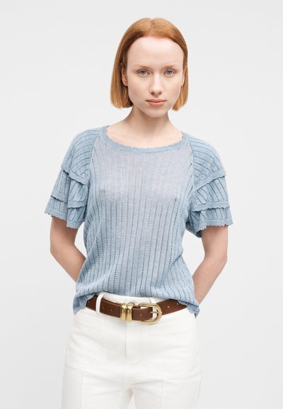 Pull en maille bleu clair avec manches courtes à volants, présentant des rayures alternées et un col rond, porté avec un pantalon blanc et une ceinture marron.