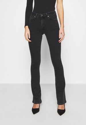 Frau trägt schwarze eng anliegende Jeans, ein schwarzes, einseitig schulterfreies Oberteil und schwarze spitze Pumps, steht vor einem weißen Hintergrund.