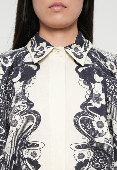 Bluză albă cu imprimeu floral bleumarin, având guler în vârf, închidere cu butoane și accente texturate. Designul include modele învârtite și detalii.
