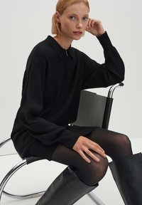 Maglione nero oversize con collo a zip, indossato sopra collant neri e stivali neri al ginocchio, seduto su una sedia moderna in metallo su uno sfondo semplice.