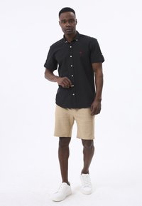 Schwarzes Kurzarmhemd mit Knopfleiste und rotem Logo, kombiniert mit beigen Shorts und weißen Turnschuhen vor einem einfarbigen Hintergrund.