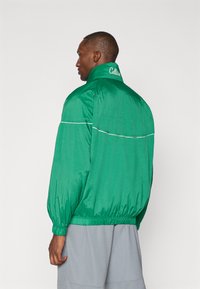 Grøn vindjakke med høj krave, elastiske manchetter og en subtil hvid stribe detalje. "Celtics" logo på den bageste krave.