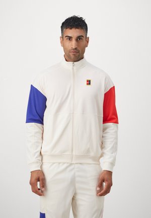 Chaqueta de entrenamiento blanca con cremallera y mangas en bloques de color azul y rojo. Textura suave con puños acanalados y un pequeño logo en el pecho.