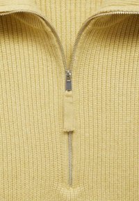 Haut en maille côtelée jaune avec une fermeture éclair centrale argentée, tissu texturé et finition douce. La fermeture éclair remonte jusqu'au col haut.