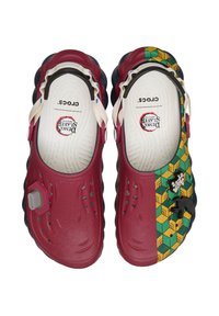 Crocs klompen met een kastanjebruine basis, waarvan de ene helft een groen en gele geometrische patroon heeft en versieringen, witte verstelbare banden.