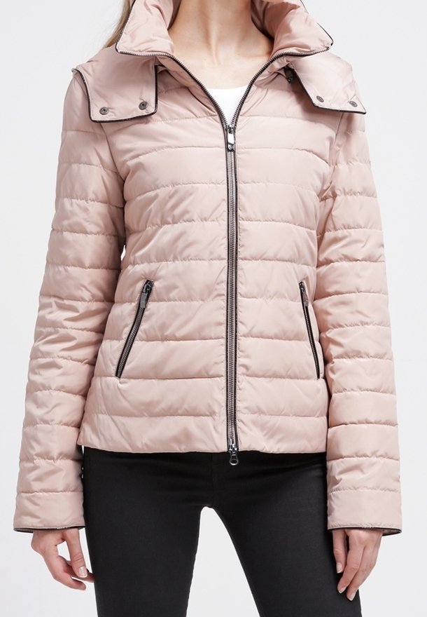 Kvinna iklädd en ljusrosa quiltad pufferjacka med svarta dragkedjefickor och hög krage, över svarta byxor.