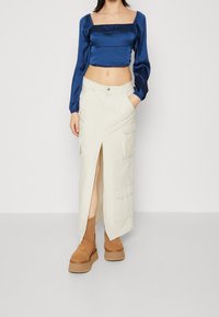 Modrý saténový crop top s nafouknutými rukávy a čtvercovým výstřihem v kombinaci s krémovou sukní s vysokým rozparkem a hnědými platformovými botami.