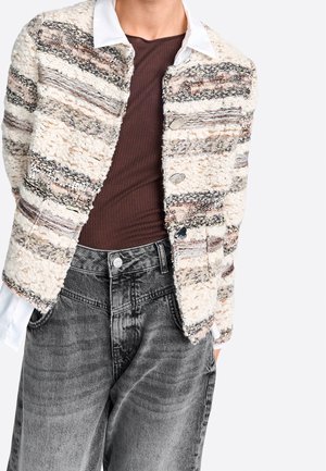 Persona che indossa una giacca a righe beige e marrone con una trama sopra una maglia marrone scuro con colletto bianco e jeans neri sbiaditi a vita alta.