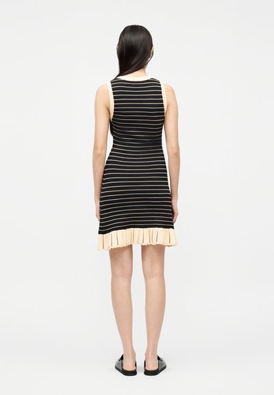 Frau mit langem, dunklem Haar, die ein ärmelloses, schwarz-beiges Kleid mit horizontalen Streifen trägt, das einen beigen, gerafften Saum und schwarze Slipper besitzt.