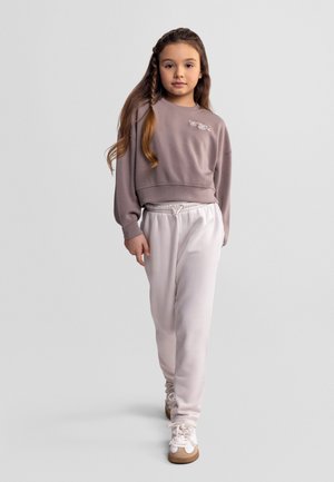 Jeune fille aux longs cheveux tressés marchant vers l'avant, portant un sweat-shirt court mauve, un pantalon de survêtement blanc et des baskets blanches.