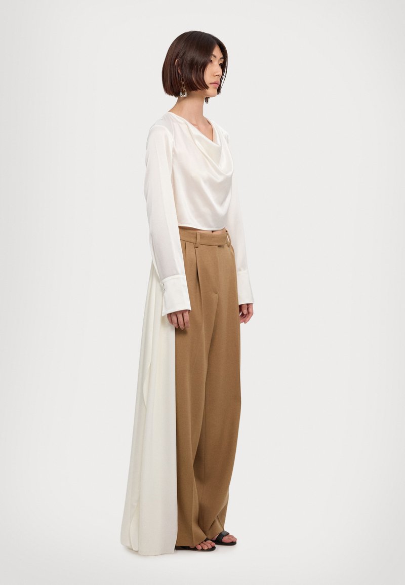 Λευκή μπλούζα με γιακά καπακιού και μακριά μανίκια συνδυασμένη με καφέ παντελόνι ψηλής μέσης και φαρδιάς γραμμής. Λείο υλικό, μινιμαλιστικός σχεδιασμός και τονική παραλλαγή χρωμάτων.