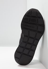 Svart idrottssko sula med texturerad gummi, med ett mönstrat däck och präglad logodetalj, vinklad mot en ljus bakgrund.