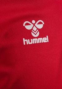 Rode mesh stof met een wit Hummel-logo. Het ontwerp bevat een symmetrisch embleem met afgeronde accenten en een centraal motief.