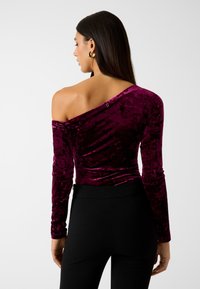 Violetter Samt-Top mit einem Schulterträger, langen Ärmeln und schmaler Passform. Mit dezentem Logo-Detail nahe der Schulter, kombiniert mit schwarzen Hosen.