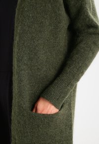 Cardigan long, vert mousse, fabriqué en tricot duveteux. Il présente une coupe décontractée, des poignets côtelés et deux poches avant pour plus de praticité.