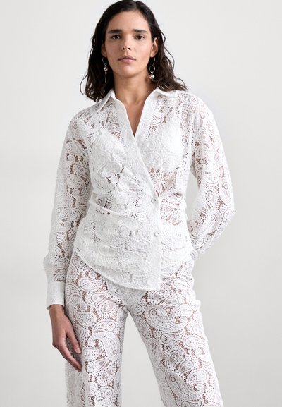 Blouse en dentelle blanche avec un motif paisley complexe, manches longues, fermeture à boutons, design superposé, assortie avec un pantalon en dentelle.