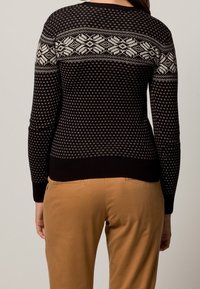 Svart stickad tröja med vita geometriska mönster på axlarna och ärmarna, med en figursydd design och ribbad nederkant. Kombination med beige byxor.