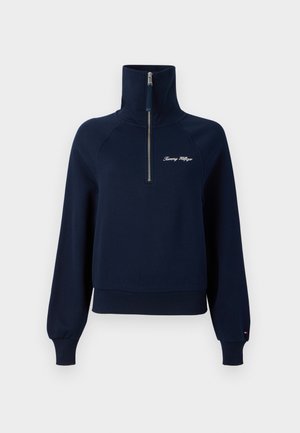Tumedat sinine pooltõmbluku ja pehme materjaliga pullover, millel on kõrge kaelus, soonikud varrastes ja rinnal brodeeritud logo.