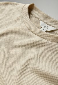 Beige tekstureret sweater med et ribmønster. Rund halsudskæring med et mærke, der står "THE SET" og størrelse "M." Nærbillede fremhæver stofkvaliteten.