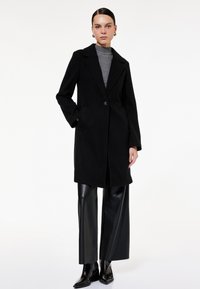 Abrigo negro de mezcla de lana con cuello de muesca, cierre de un botón y bolsillos laterales, superpuesto a un suéter de cuello alto de canalé gris y pantalones de cuero sintético.