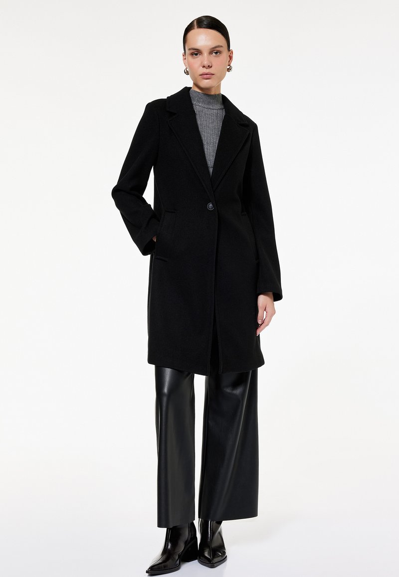 Abrigo negro de mezcla de lana con cuello de muesca, cierre de un botón y bolsillos laterales, superpuesto a un suéter de cuello alto de canalé gris y pantalones de cuero sintético.