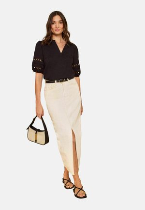 Femme portant un chemisier noir à col en V avec manches en dentelle, une jupe fendue crème taille haute, une ceinture noire, des sandales noires, tenant un sac à main beige et noir.