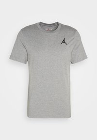 T-shirt en coton gris à manches courtes, col rond, présentant un logo Jumpman noir sur le côté gauche de la poitrine. Design simple et texture lisse.