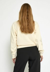 Persona con lunghi capelli castani che indossa una felpa crema con motivi sulle maniche e pantaloni neri, in piedi con le spalle alla fotocamera.