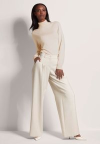 Beige coltrui gecombineerd met lichtbeige wijde broek. De broek heeft plooien en een tailleband met een enkele knoop. De schoenen zijn wit.