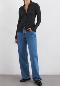 Person som bär en svart ribbad knapp-up cardigan, blå vida jeans och svarta loafers, stående mot en vit vägg.