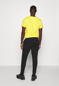 Gul bomulds t-shirt og sorte sweatpants iført med sorte sneakers, set fra ryggen, hvilket viser trøjen med rund halsudskæring og buksernes lommer.