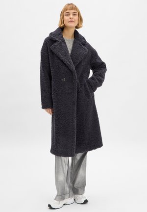 Dunkelgrau beschichteter Fleece-Mantel mit breitem Revers und doppelten Knöpfen. Kombiniert mit glänzenden silbernen Hosen und weißen Turnschuhen.
