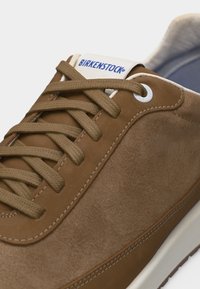 Καφέ σουέντ και δερμάτινο αθλητικό παπούτσι με ταμπά κορδόνια, λευκή σόλα και μπλε λογότυπο Birkenstock στη γλώσσα, κοντινή όψη.