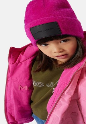 Bonnet en fausse fourrure rose avec une étiquette noire, veste imperméable rose vif, pull vert olive avec des lettres en strass, et cheveux lisses et noirs.