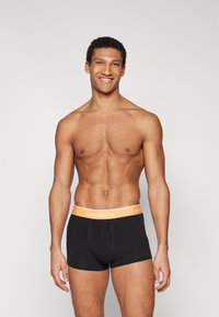 Svarta bomullskalsonger med en midjeband i orange Lacoste-logga, som erbjuder en tät passform och en mjuk yta. Modellen står självsäkert framför en neutral bakgrund.