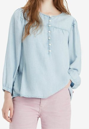 Frau trägt eine hellblaue langärmelige Bluse mit Knöpfen vorne und hellrosa Hose, Hand in der Tasche auf weißem Hintergrund.