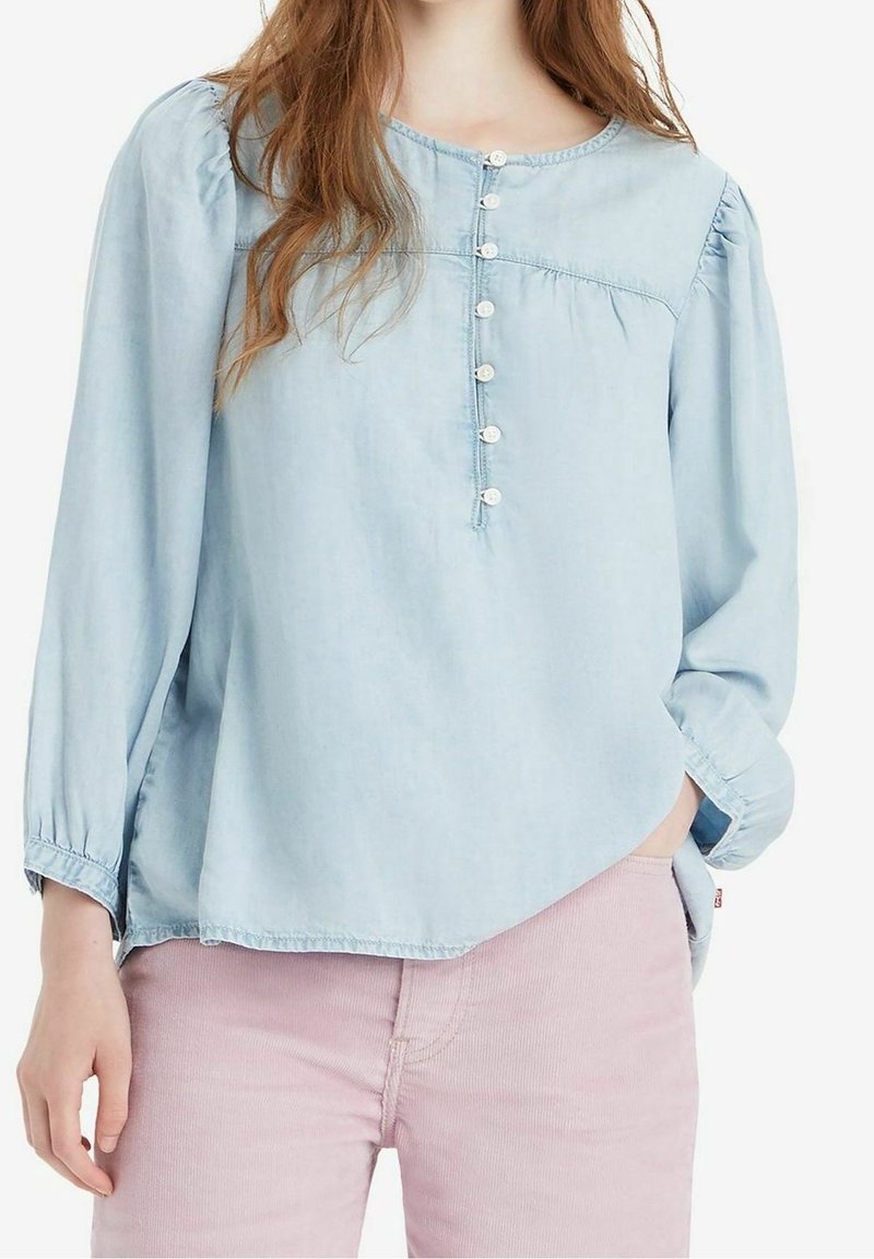 Femme portant un chemisier à manches longues bleu clair avec des boutons sur le devant et un pantalon rose clair, main dans la poche sur fond blanc.