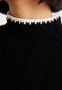 Czarny sweter z dzianiny z wysokim kołnierzem ozdobionym białym, teksturowanym kontrastowym obszyciem. Żebrowany wzór podkreśla wizualne detale i fakturę.