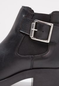 Botín de cuero negro hasta el tobillo con un acento de hebilla plateada, paneles laterales de tela elástica y un tacón grueso negro. Textura suave y líneas limpias.