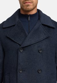 Abrigo navy de doble botonadura, confeccionado en tejido texturizado, con amplias solapas, dos botones negros y una capa interior con cremallera en el cuello.