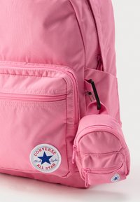 Zaino in tessuto rosa con un compartimento principale, tasca frontale con cerniera e una piccola pochette staccabile. Presenta un logo circolare "Converse All Star".