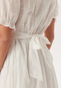 Bílá bavlněná šaty s výšivkou květinových vzorů a výřezovými detaily, s nabíranými rukávy a páskem v pase. Měkká, lehká látka.