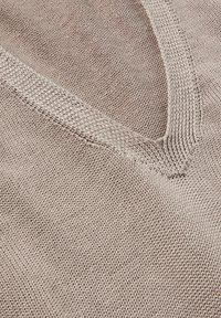 Beiger Strickstoff mit V-Ausschnitt-Design. Verfügt über ein strukturiertes Muster mit lockerem Gewebe, das eine weiche und atmungsaktive Optik erzeugt.